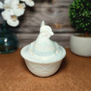 May include: A white iridescent glass hen on a nest dish featuring a detailed chicken lid with a textured feather pattern and a base with a diamond lattice design and a rope-style rim. The glass has a pearlescent finish that reflects light.
