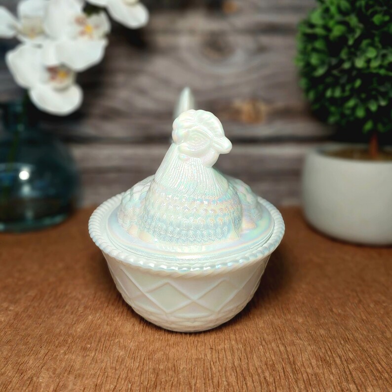 May include: A white iridescent glass hen on a nest dish featuring a detailed chicken lid with a textured feather pattern and a base with a diamond lattice design and a rope-style rim. The glass has a pearlescent finish that reflects light.