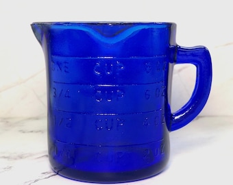 May include: A cobalt blue glass measuring cup with a handle and a pouring spout. The exterior features embossed measurement markings in cups and ounces, including 1/4 cup, 1/2 cup, 3/4 cup, and one cup, along with corresponding 2 oz, 4 oz, 6 oz, and 8 oz labels.