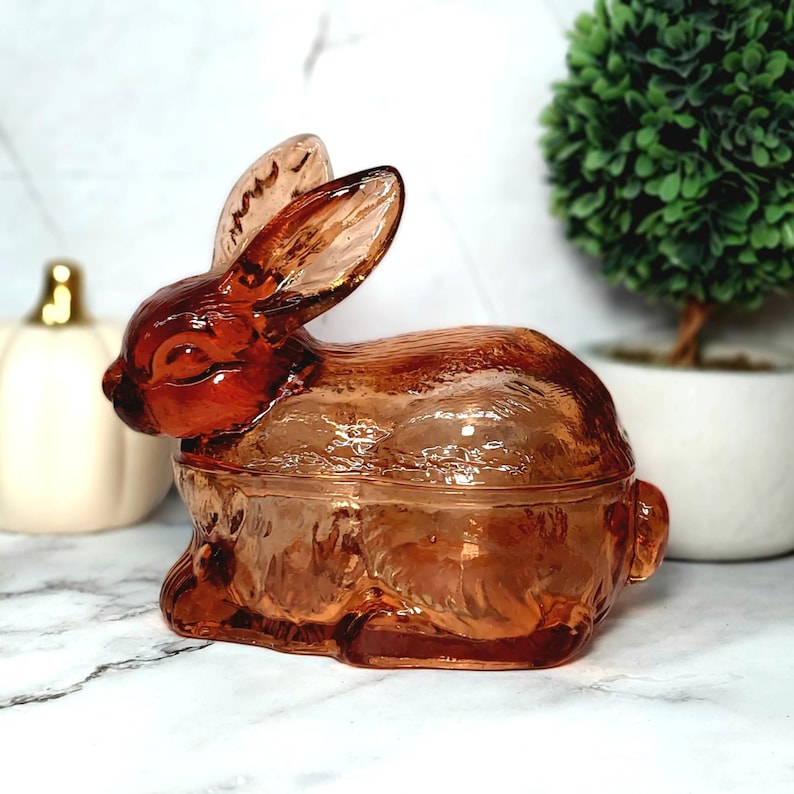 Op de afbeelding: Een decoratieve glazen konijnvormige sieradendoos in een warme amberkleur. Het konijn heeft lange oren en zit rechtop. De doos wordt waarschijnlijk gebruikt voor het opbergen van kleine items. Het glas heeft een getextureerd uiterlijk.