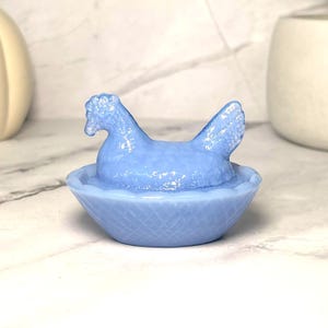 May include: A light blue glass hen figurine in a basket-shaped dish. The hen is sitting in the basket, with its head and tail visible. The basket has a woven texture. This decorative piece is suitable for home decor.