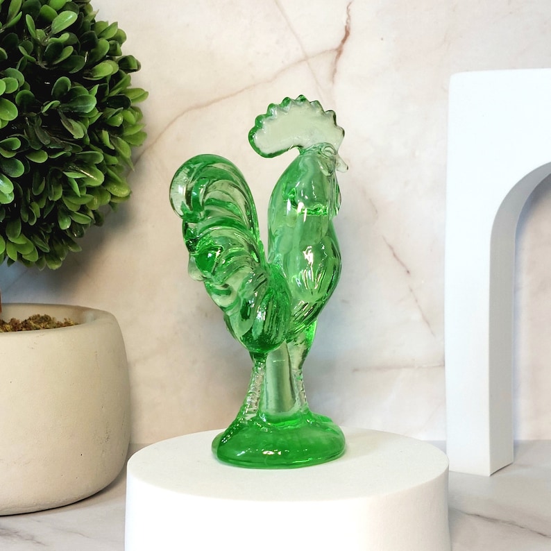 May include: A green glass rooster figurine with a detailed design. The rooster is standing on a pedestal base.