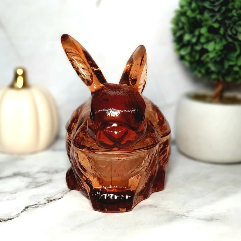 Op de afbeelding: Een decoratieve, amberkleurige glazen sieradendoos in de vorm van een konijn. Het konijn heeft lange oren en een gedetailleerd gezicht. De doos staat op een wit oppervlak met een pompoen en een kleine plant in een pot op de achtergrond.