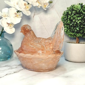 May include: A decorative glass hen figurine in a nest. The hen is a translucent peach color, with detailed feather and wing textures. The nest is also peach-colored, with a woven basket design. The figurine is a unique home decor item.