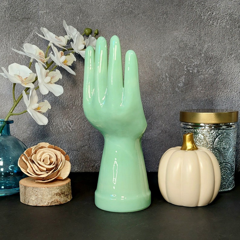 May include: A ceramic hand sculpture in a light green color. The hand is raised with fingers spread apart. The sculpture is on a table with other decorative items.
