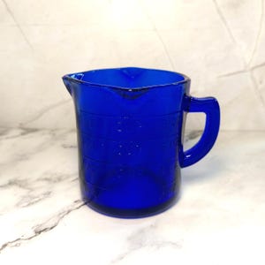 May include: A cobalt blue glass measuring cup featuring embossed measurement markings in cups and ounces. The vessel includes a pouring spout and a sturdy side handle, with clear, raised text indicating increments of 1/4, 1/2, 3/4, and 1 cup, as well as 2, 4, 6, and 8 ounces.