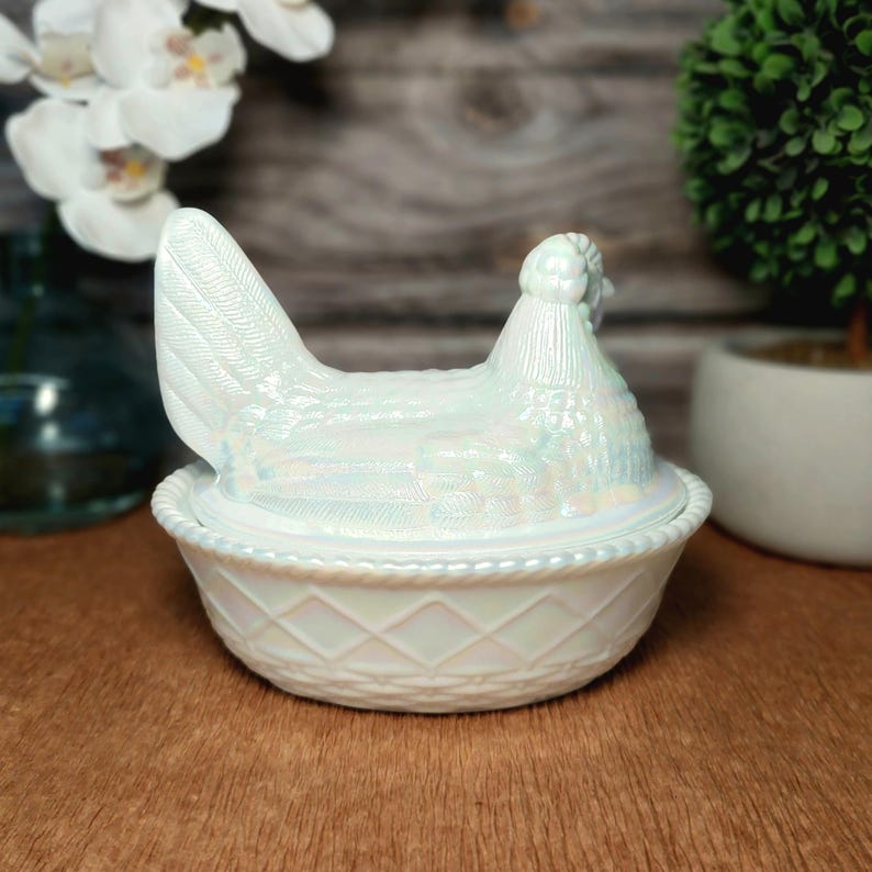 May include: An iridescent white glass hen on a nest dish. The lid features a moulded hen with detailed feather textures, while the base is shaped like a woven basket with a diamond pattern and a rope-style rim. The glass has a pearlescent finish.