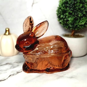 May include: A decorative, amber-colored glass bunny-shaped trinket box. The bunny has long ears and is sitting upright. The box is a light amber color and has a lid. The background includes a white pumpkin and a small green plant.