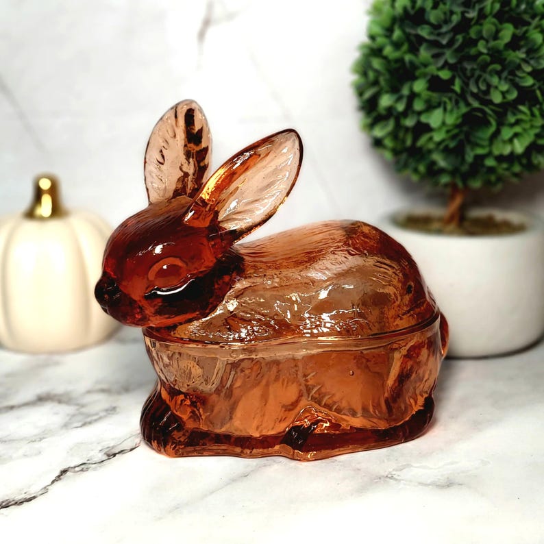 Op de afbeelding: Een decoratieve, amberkleurige glazen konijnvormige sieradendoos. Het konijn heeft lange oren en zit rechtop. De doos heeft een licht amberkleur en heeft een deksel. De achtergrond bevat een witte pompoen en een kleine groene plant.