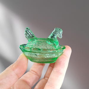 May include: A green glass chicken dish with a textured surface. The chicken is sitting in a nest and has its wings spread out.