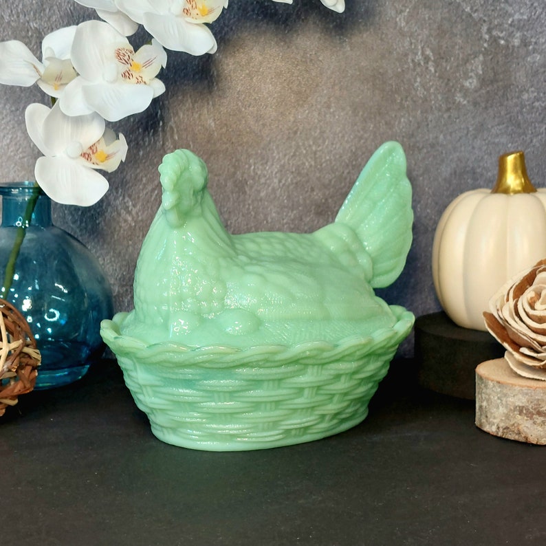 May include: A light green hen-shaped dish with a basket weave base. The hen is sitting on the basket, and the dish is made of glass. The dish is a decorative item, and it is likely used for storing small items or as a decorative piece.