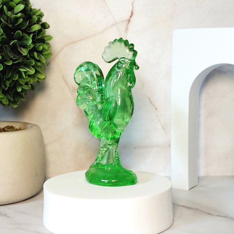 May include: A green glass rooster figurine with a detailed design. The rooster is standing on a white pedestal.