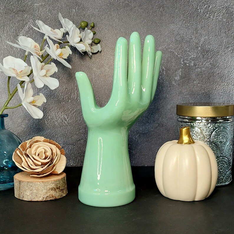 May include: A ceramic hand sculpture in a light green color. The hand is raised with fingers extended, resembling a wave. The sculpture is on a table with a white pumpkin and a glass jar with a gold lid.