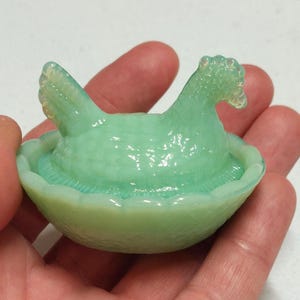 May include: A green glass chicken and nest dish. The chicken is sitting in a nest with a ruffled edge.