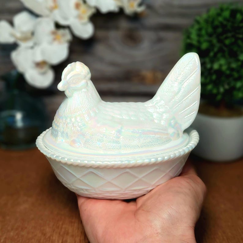 May include: A white iridescent glass hen on a nest dish. The lid is shaped like a sitting hen with textured feather details, while the base features a raised diamond lattice pattern and a beaded rim, creating a reflective, pearlescent finish.