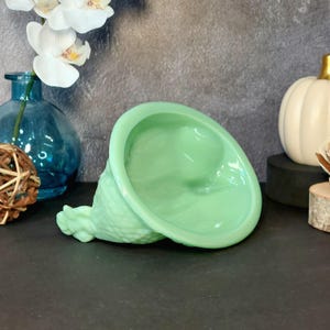 May include: A light green, cone-shaped decorative object with a textured base and a smooth, bowl-like top. The object is lying on its side, showcasing its unique form. The background includes a blue vase, white flowers, and a white pumpkin.