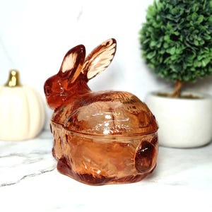 Op de afbeelding: Een decoratieve glazen konijnvormige container in een warme amberkleur. Het konijn heeft lange oren en een getextureerd lichaam, met een deksel dat goed past. De container wordt waarschijnlijk gebruikt voor het opbergen van kleine items of als decoratief stuk.