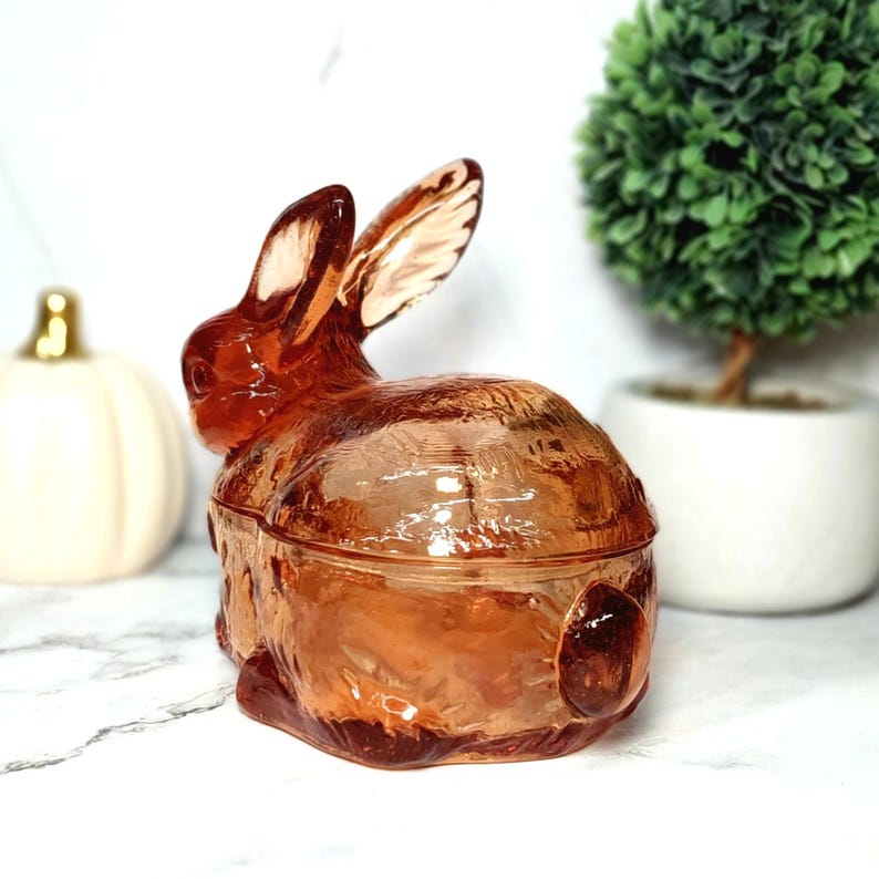 Op de afbeelding: Een decoratieve glazen konijnvormige container in een warme amberkleur. Het konijn heeft lange oren en een getextureerd lichaam, met een deksel dat goed past. De container wordt waarschijnlijk gebruikt voor het opbergen van kleine items of als decoratief stuk.