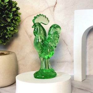 May include: A green glass rooster figurine with a detailed design. The rooster has a raised crest and a long tail.