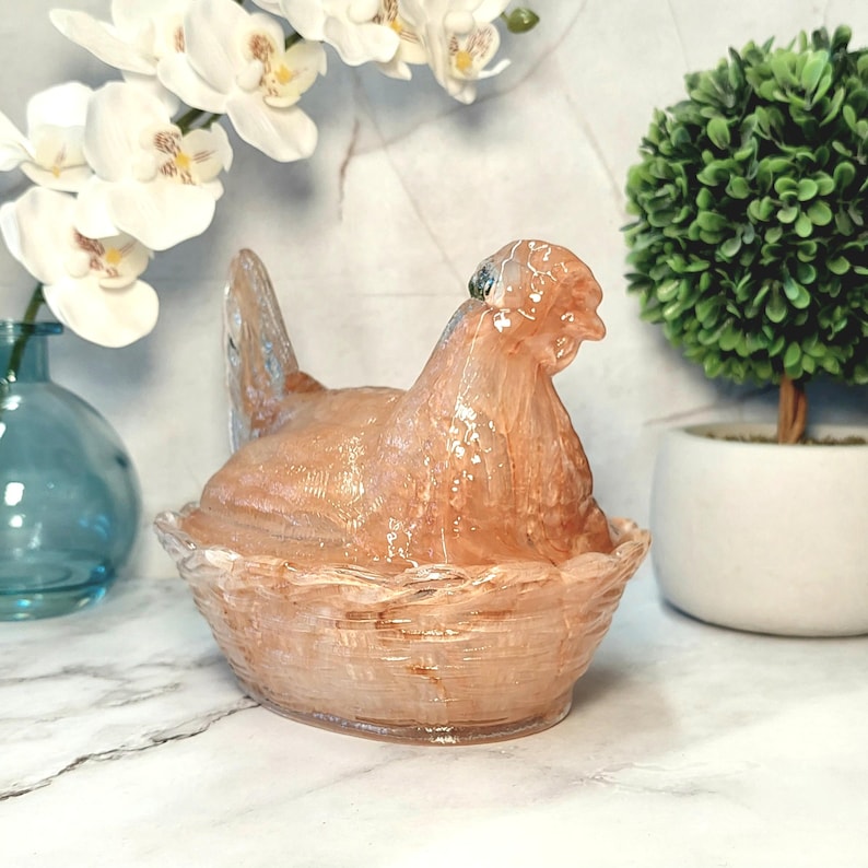 May include: A decorative glass hen figurine in a basket-like base. The hen is a translucent peach color, and the basket is a similar shade. The figurine is a unique home decor item.