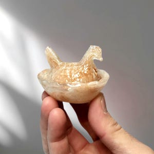 May include: A translucent, amber-colored glass sculpture of a hen sitting in a nest. The hen has a detailed, textured surface, and the nest is a shallow bowl shape. The sculpture is held in a hand against a neutral background.