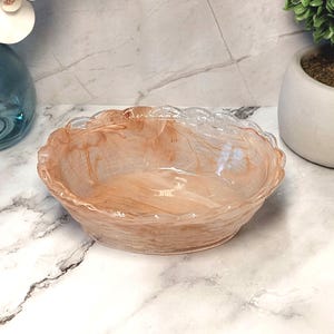 May include: A decorative, oval-shaped bowl with a scalloped rim. The bowl features a marbled design in shades of peach and cream, creating a translucent effect. The bowl is likely made of glass or a similar material, and is suitable for decorative purposes.