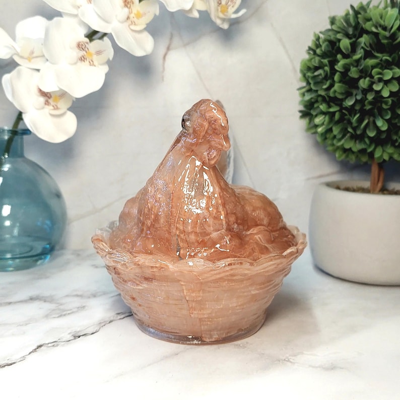May include: A translucent, peach-colored decorative bowl with a lid. The bowl has a textured, woven-like design around the base and lid. The lid is shaped like a hen. The bowl is sitting on a white surface.