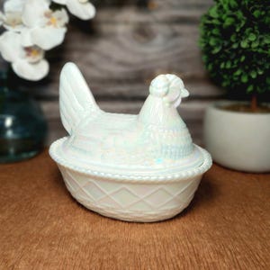 May include: An iridescent white glass hen on a nest dish featuring a detailed chicken figurine lid resting on a base with a moulded diamond lattice pattern and a rope-textured rim. The glass has a pearlescent finish that reflects light.