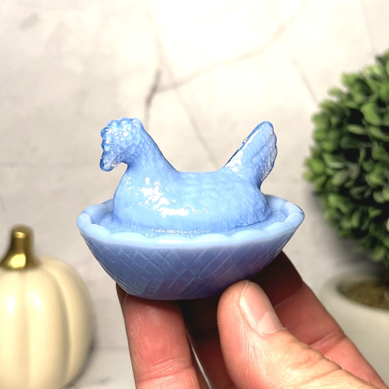 May include: A light blue hen figurine in a basket. The hen is sitting in a woven basket. The figurine is made of glass or ceramic. The hen has a detailed comb and tail feathers. The basket has a textured pattern.