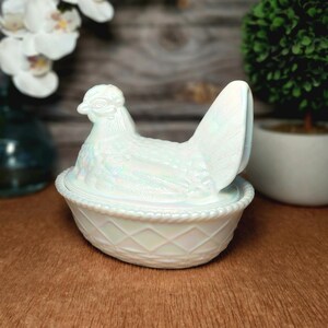 May include: An iridescent white glass hen on a nest dish. The lid is shaped like a sitting hen with detailed feather textures, while the base features a raised diamond lattice pattern and a rope-style rim, creating a shimmering, pearlescent finish.
