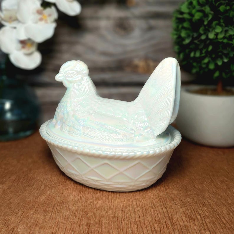May include: An iridescent white glass hen on a nest dish. The lid is shaped like a sitting hen with detailed feather textures, while the base features a raised diamond lattice pattern and a rope-style rim, creating a shimmering, pearlescent finish.
