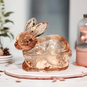 May include: A pink glass rabbit shaped candy dish with a lid. The dish is sitting on a wooden surface.
