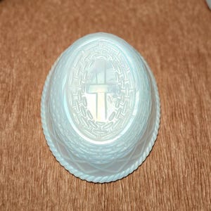 May include: An oval-shaped, iridescent white glass dish featuring a central embossed cross surrounded by a woven basket-weave pattern. The rim of the dish is decorated with a twisted rope-like texture, and the base has a diamond-shaped pattern.