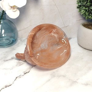 May include: A decorative, round, marbled resin dish in shades of peach and white. The dish has a small, textured base and a glossy finish. The product is suitable for holding small items.