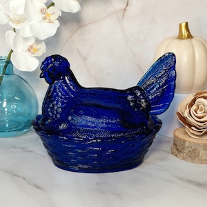 May include: A cobalt blue glass hen sitting in a nest. The hen is a vintage style butter dish.