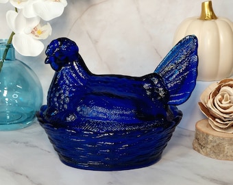 May include: A cobalt blue glass hen on a nest-shaped base. The decorative piece has a textured surface and is likely used for display or storage. The hen is perched atop a woven basket design.