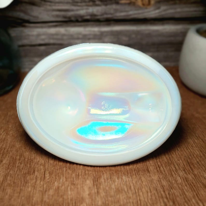 May include: An oval-shaped white glass dish featuring a smooth, iridescent finish that reflects light with subtle rainbow hues. The centre of the dish displays a small, embossed circular maker's mark, and the edges are slightly raised and rounded.