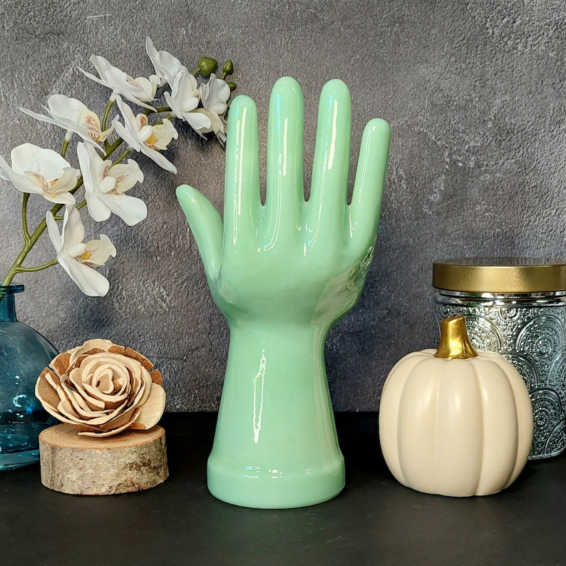 May include: A light green ceramic hand sculpture with five fingers. The hand is raised with the palm facing outward. The sculpture is on a table with a white pumpkin and a glass jar in the background.