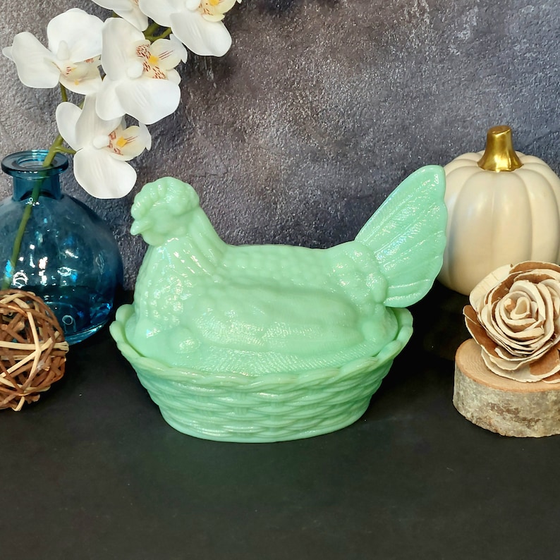 May include: A light green hen-shaped trinket box. The hen sits atop a basket-weave base. The box is made of glass or ceramic. The background includes a blue vase, white flowers, and a white pumpkin with a gold stem.