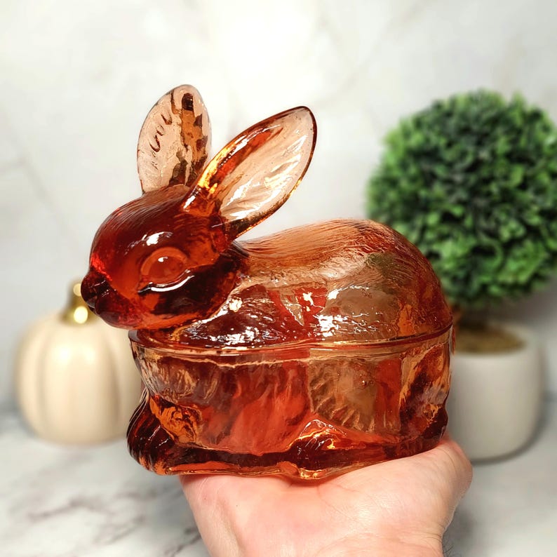 Op de afbeelding: Een amberkleurige glazen konijnvormige sieradendoos. Het konijn heeft grote oren en een gedetailleerd gestructureerd oppervlak. Het deksel past goed op de basis en cre&euml;ert een decoratief en functioneel stuk.