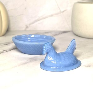 May include: A light blue, hen-shaped trinket box with a matching round bowl. The hen lid has textured details and is open, revealing the bowl. The bowl has a woven pattern and a scalloped edge. The set is made of glass.