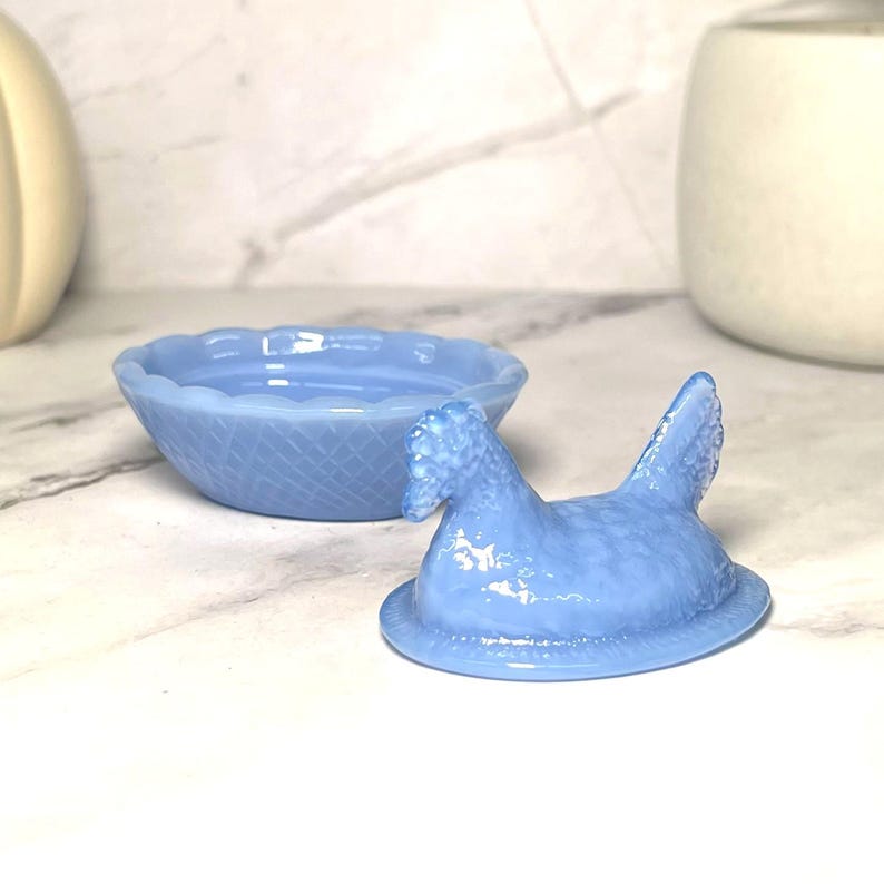 May include: A light blue, hen-shaped trinket box with a matching round bowl. The hen lid has textured details and is open, revealing the bowl. The bowl has a woven pattern and a scalloped edge. The set is made of glass.