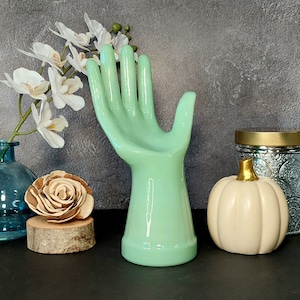 May include: A light green ceramic hand sculpture with a smooth, glossy finish. The hand is posed with fingers extended and the palm facing up. The sculpture is a decorative piece for a home or office.