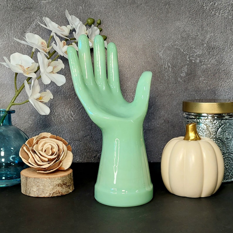 May include: A light green ceramic hand sculpture with a smooth, glossy finish. The hand is posed with fingers extended and the palm facing up. The sculpture is a decorative piece for a home or office.