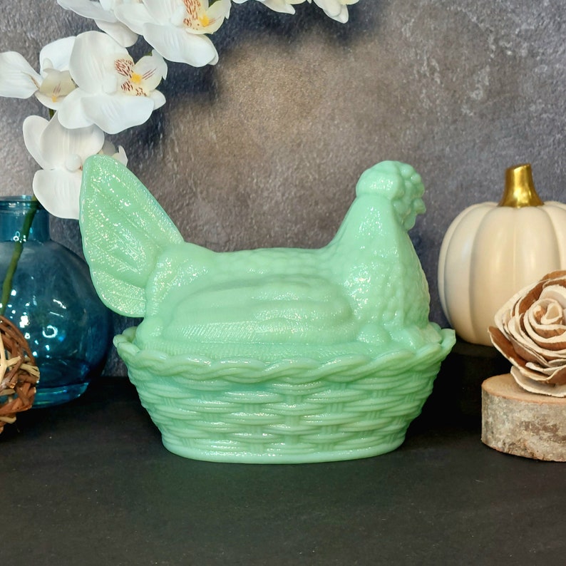 May include: A light green hen-shaped dish with a basket-weave base. The hen sits atop the basket, with detailed feather and head textures. The dish is a pastel green color, and is likely made of glass or ceramic.