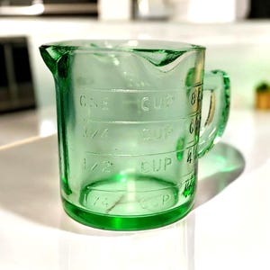 May include: A vintage green glass measuring cup with a handle and spout. The cup has measurements for 1/4, 1/2, 3/4, and 1 cup. The cup is made of clear glass and is sitting on a white surface.