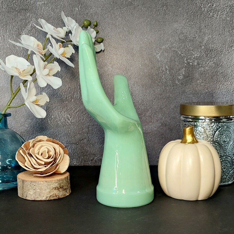 May include: A ceramic hand-shaped ring holder in a light green color. The holder is standing on a table with a white pumpkin and a decorative jar in the background.