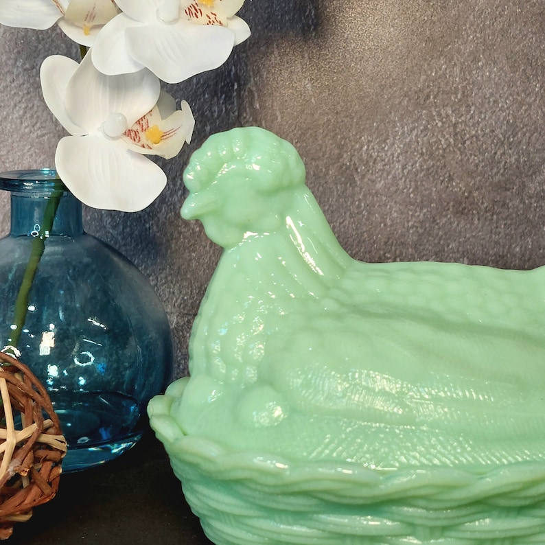 May include: A light green hen-shaped dish with a textured surface. The dish is made of glass and has a basket-weave base. A blue glass vase and white orchid flowers are in the background.