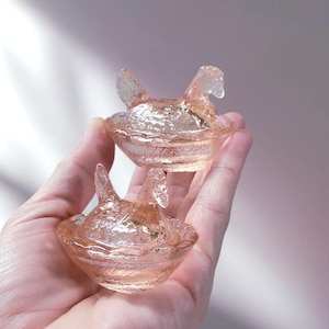 May include: Two small, pink glass hen-shaped dishes. Each hen sits in a shallow bowl, with textured details on the glass. The dishes are held in a hand, showcasing their delicate design and color.