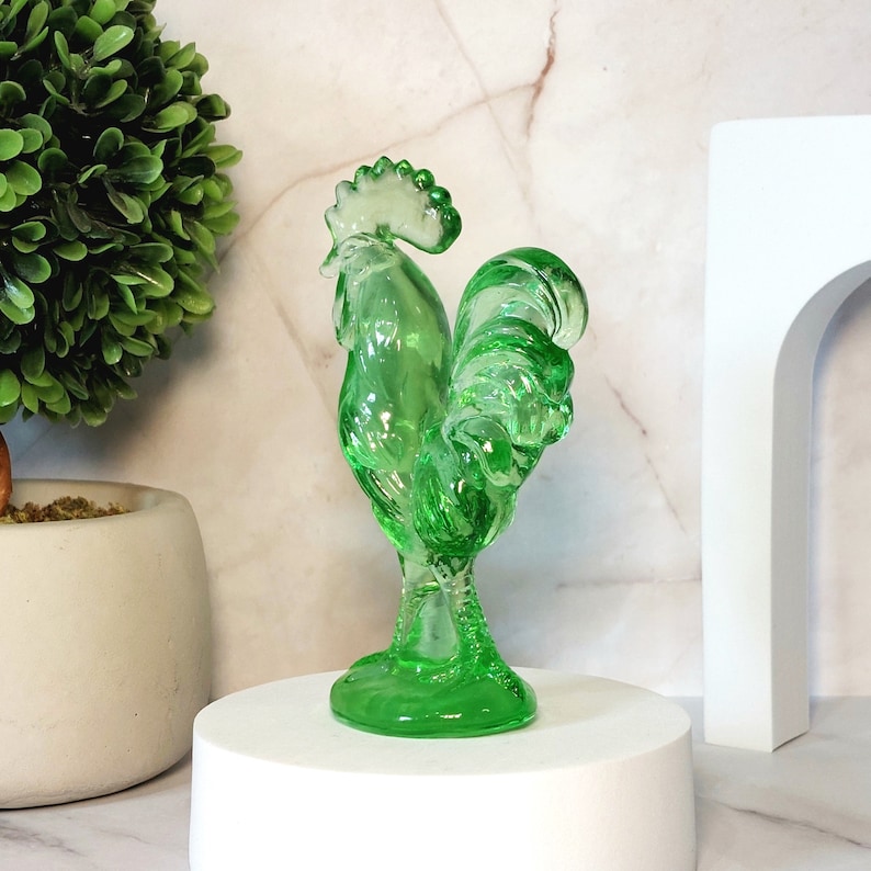 May include: A green glass rooster figurine with a detailed design. The rooster is standing on a white pedestal.